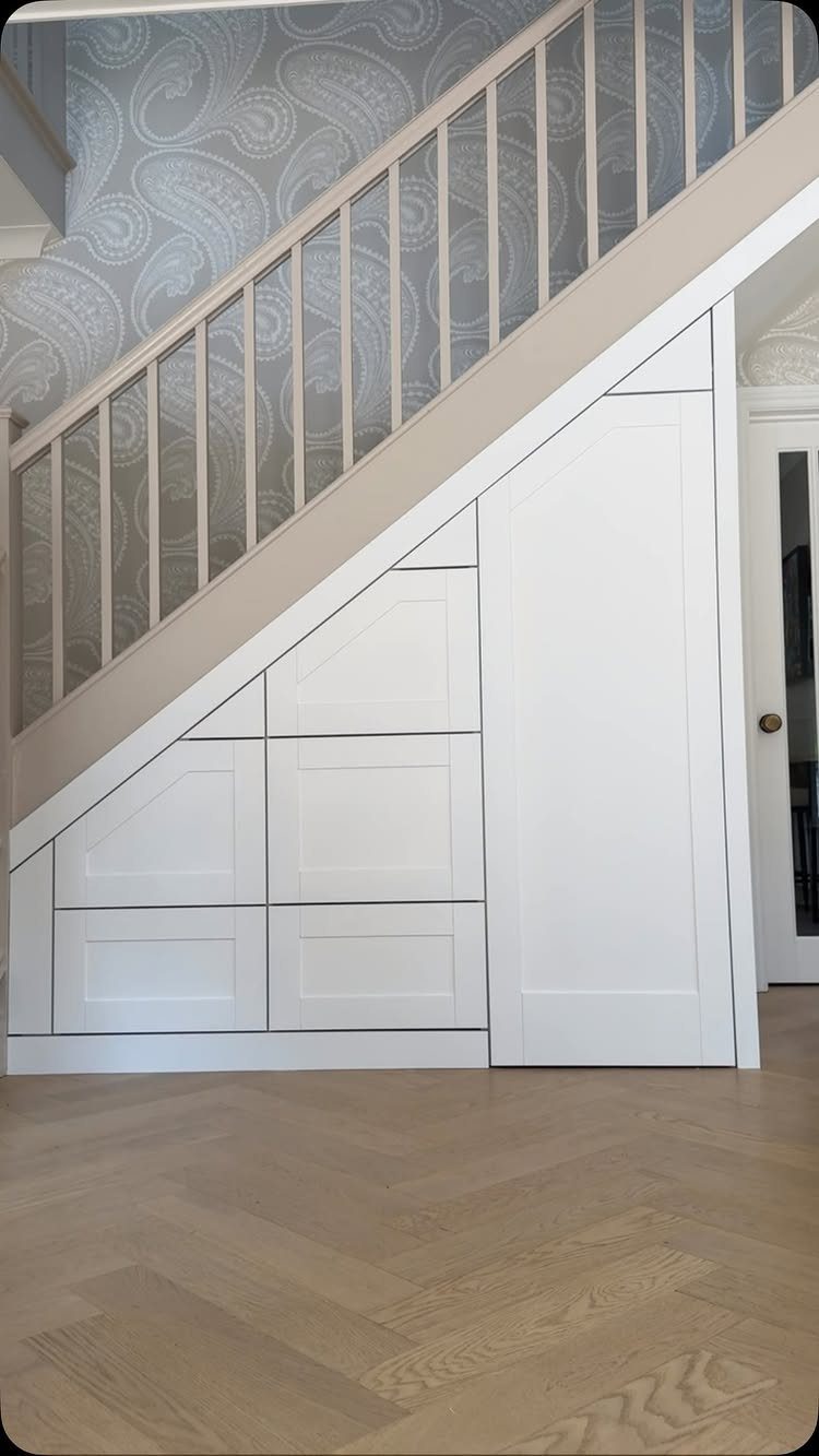 White built-in storage cabinets installed underneath a wooden staircase with paisley wallpaper and herringbone flooring.