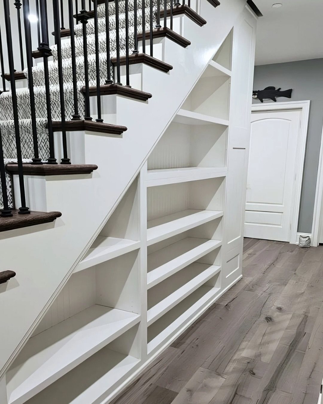 White built-in storage shelves installed underneath a dark wood staircase in a modern basement.