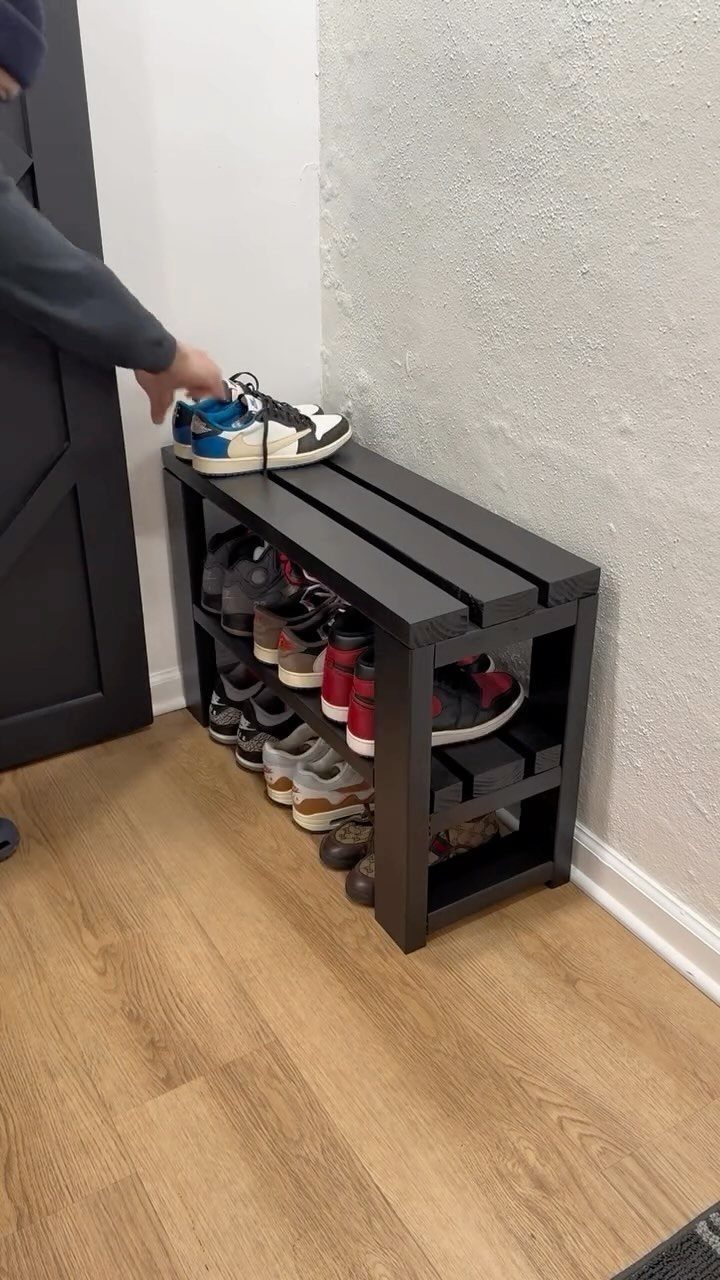 A person placing sneakers on a dark grey wooden shoe shelf bench against a white wall.