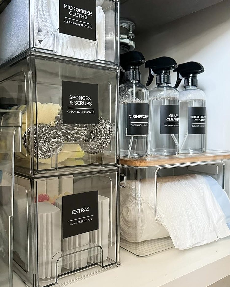 Clear stackable plastic bins filled with cleaning supplies sit on a white shelf next to spray bottles.
