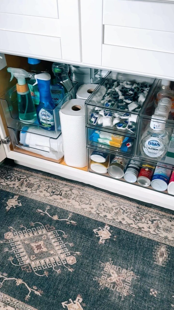Cleaning supplies organized in clear plastic bins inside a white kitchen cabinet under a counter.
