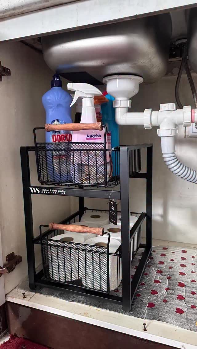 A black two-tier wire organizer holding cleaning spray bottles and toilet paper rolls under a kitchen sink.