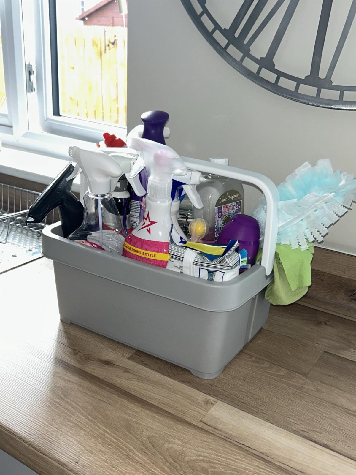 A grey plastic cleaning caddy filled with spray bottles and cloths on a wooden counter.