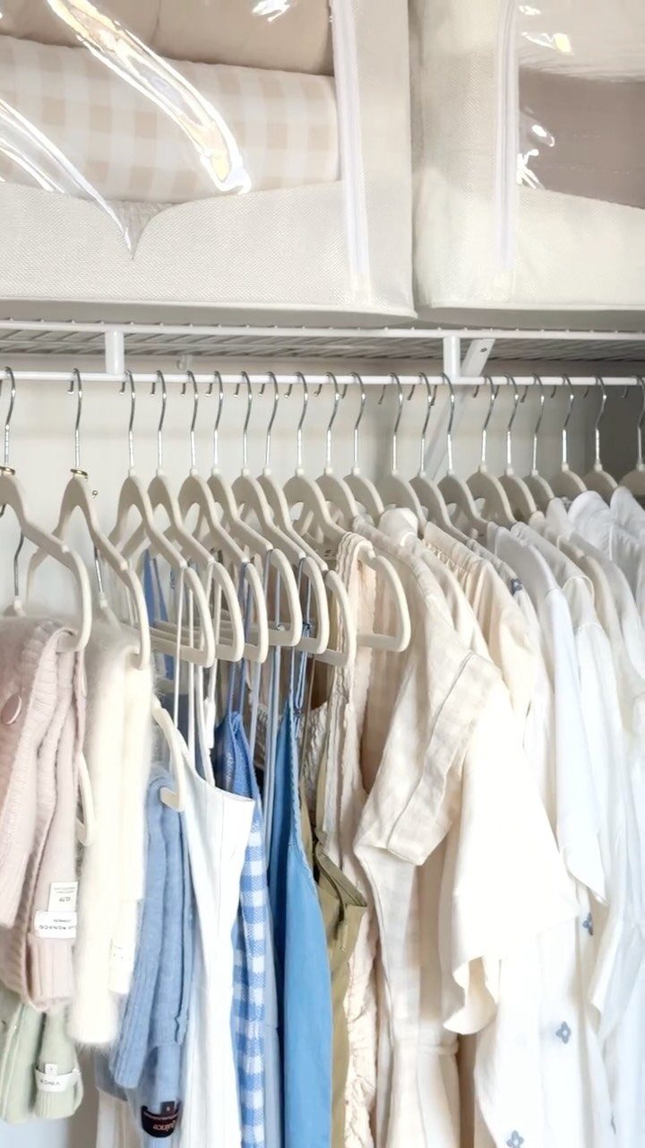 White clothes hanging on thin hangers beneath large beige fabric storage bins on a white shelf.