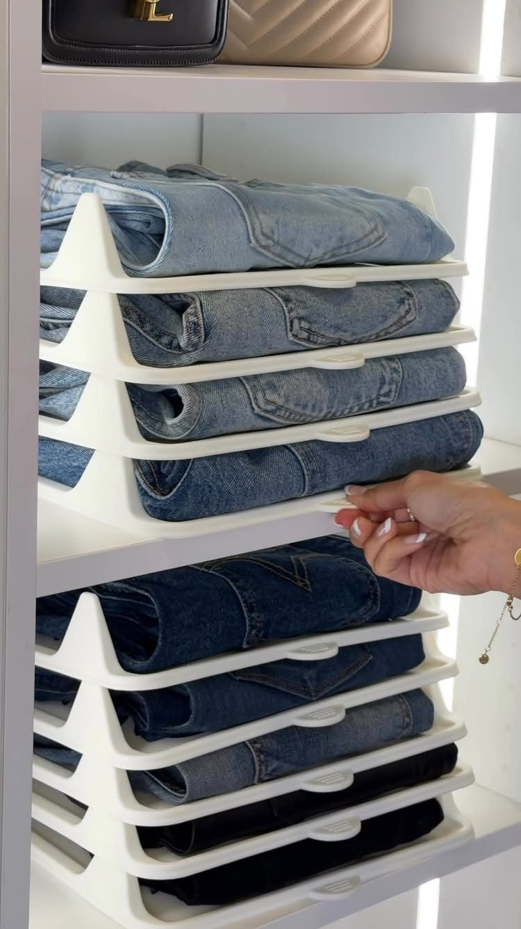 A hand reaches for folded blue jeans organized in white tiered shelf dividers within a closet.