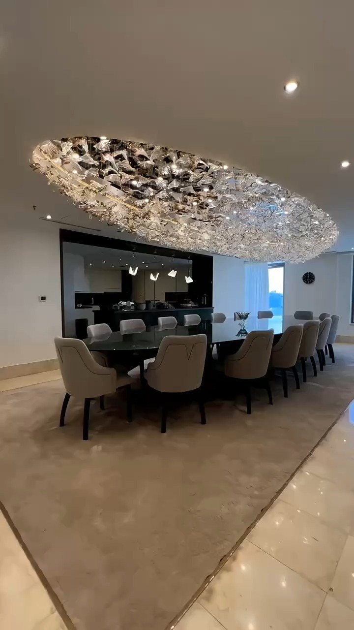A massive oval crystal chandelier hanging above a contemporary dining table and grey chairs.