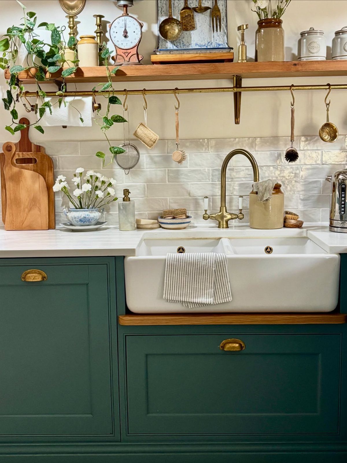 Green kitchen cabinets with a white farmhouse sink, brass faucet, and wooden shelving against subway tiles.