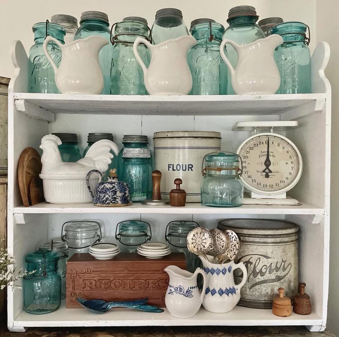 Vintage glass jars, ceramic pitchers, and antique kitchen canisters displayed on white wooden shelves.