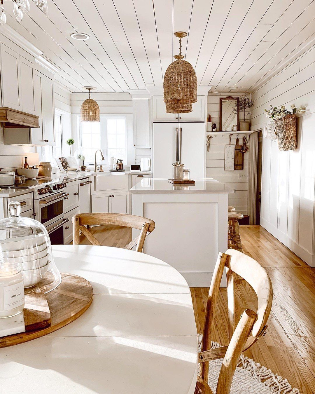 White farmhouse kitchen with shiplap walls, white cabinets, and wooden furniture in bright sunlight.