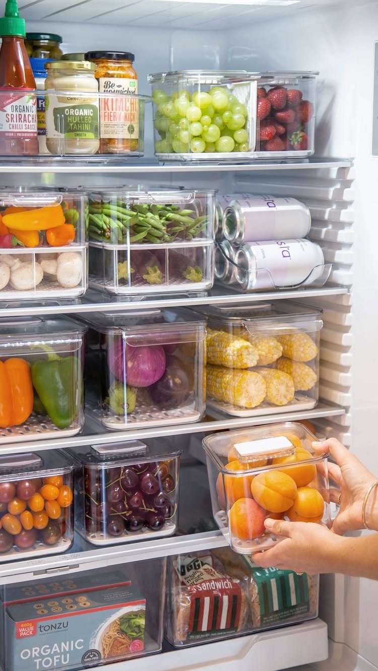 Organized refrigerator shelves filled with clear containers of fresh fruits and vegetables being arranged by hands.