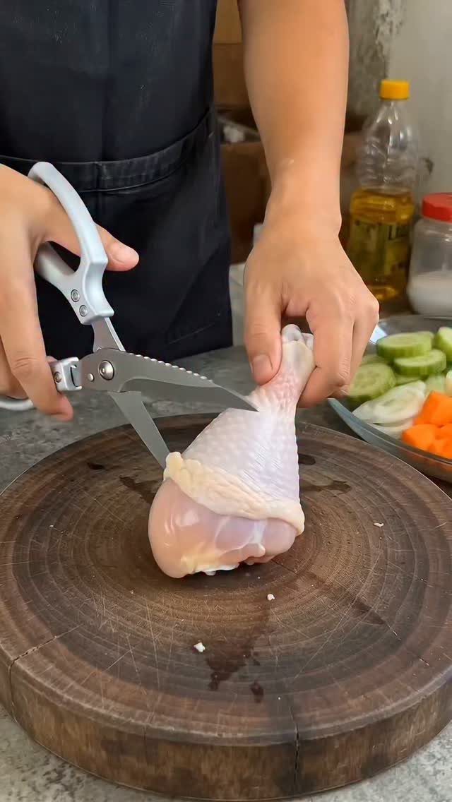 Hands using silver kitchen shears to cut a raw chicken leg on a wooden cutting board.