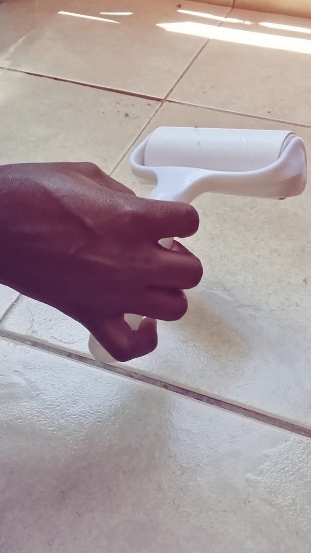 A hand using a white lint roller on a light-colored tiled floor surface.