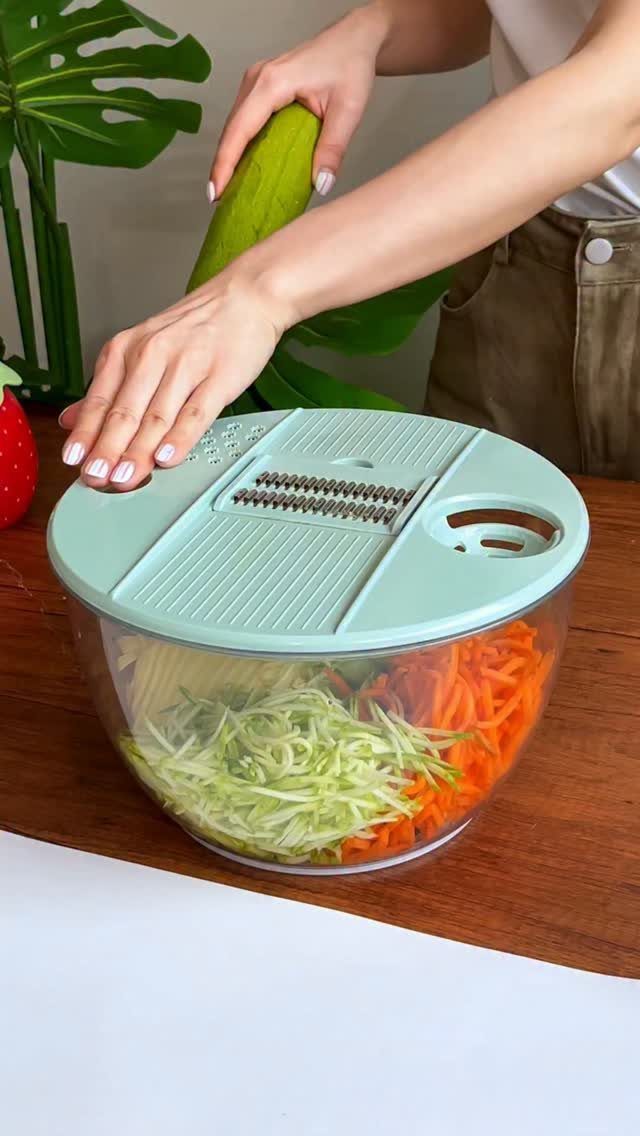 Hands using a light green manual slicer to shred a cucumber into a clear bowl filled with vegetables.