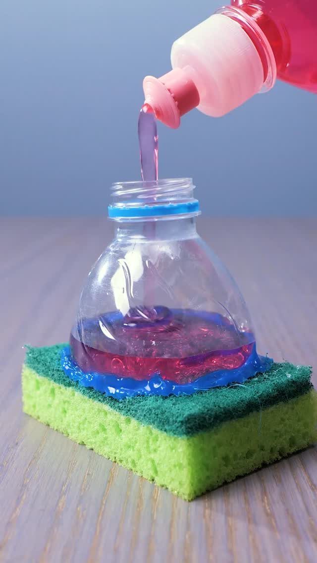 Pink liquid soap being poured into a cut plastic bottle resting on a green sponge.