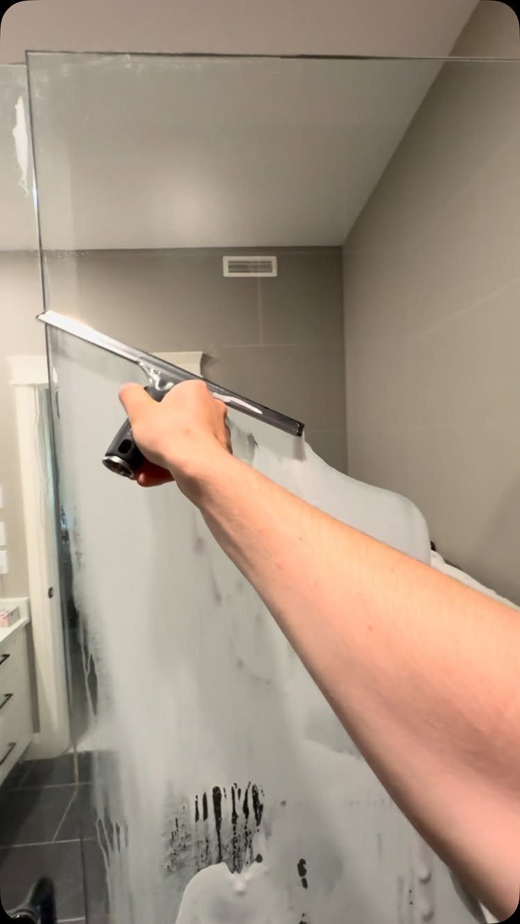 A hand using a squeegee to clean soap suds off a glass shower door.