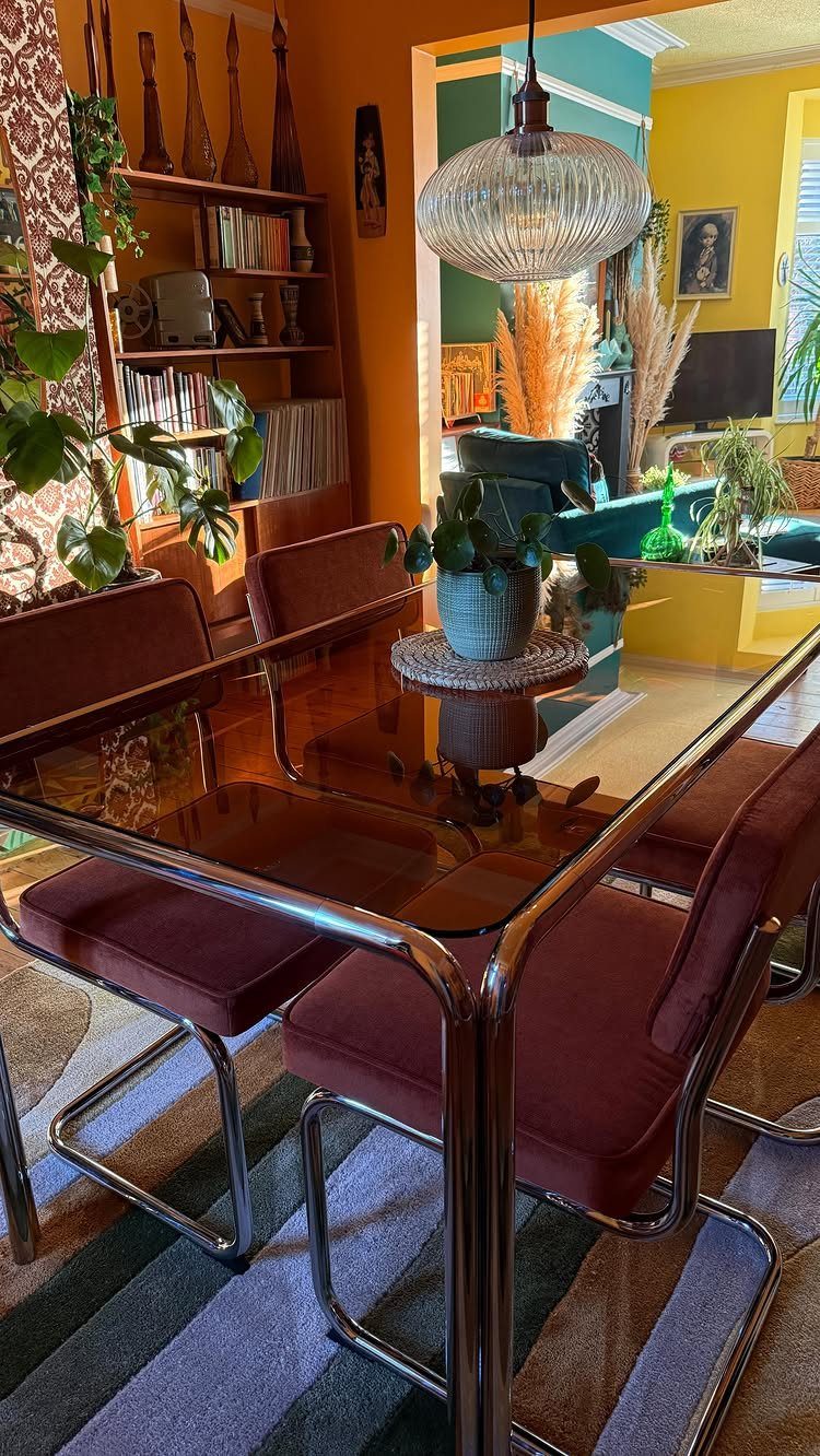 A glass dining table with maroon velvet chairs sits on a striped rug in an eclectic room.