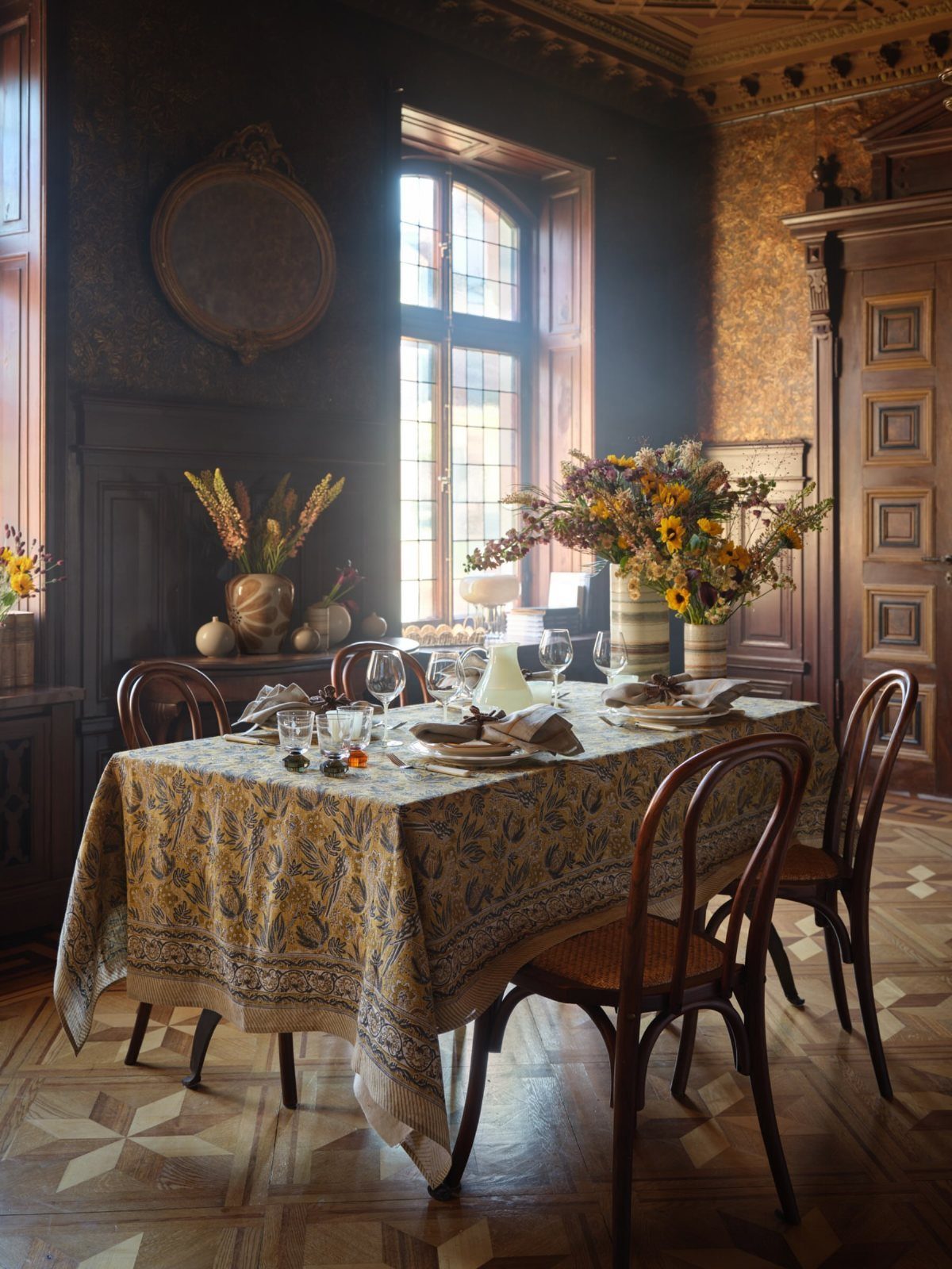 A dining table set with a patterned tablecloth, glassware, and flowers in a dark, ornate room.