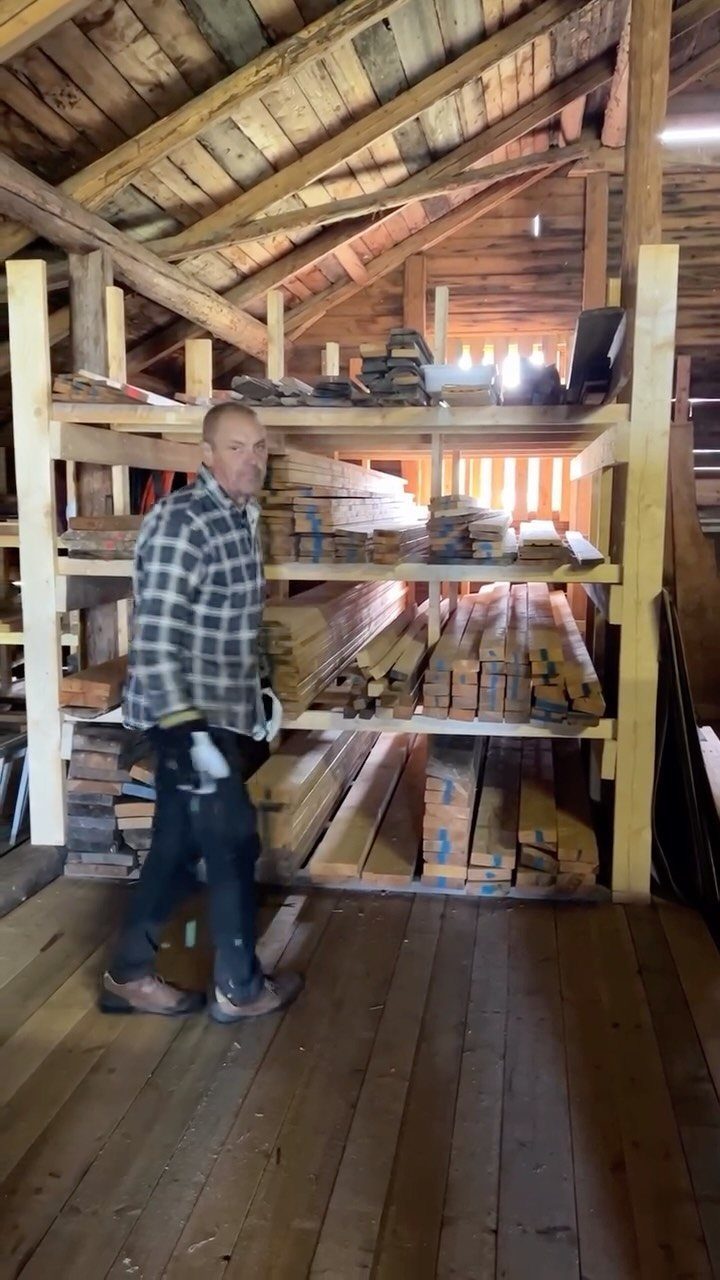 A man walking past large wooden shelves filled with various pieces of lumber in a barn.