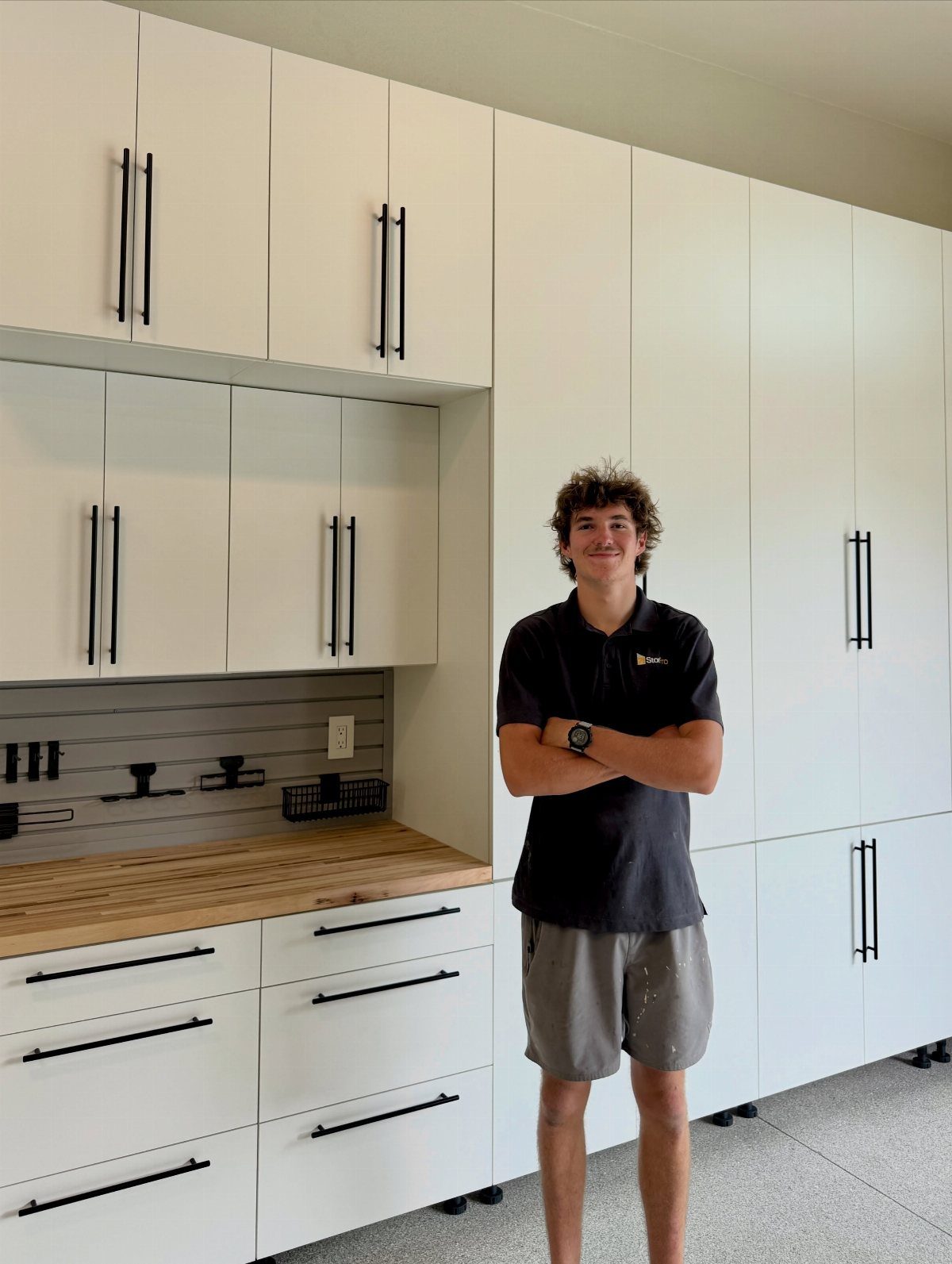 White garage cabinets with black handles and a wooden countertop against a grey slatwall.