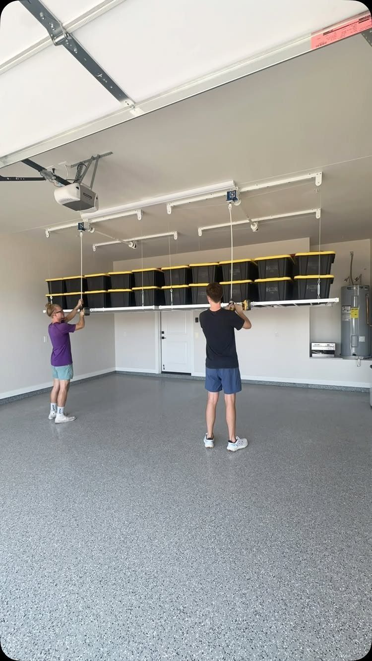 Two people adjusting a ceiling-mounted storage system with black and yellow bins in a garage.