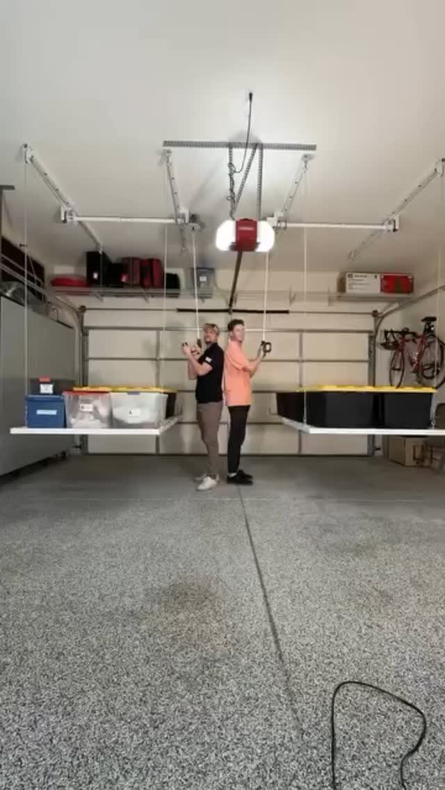 Two people using a pulley system to lift storage platforms filled with bins in a garage.
