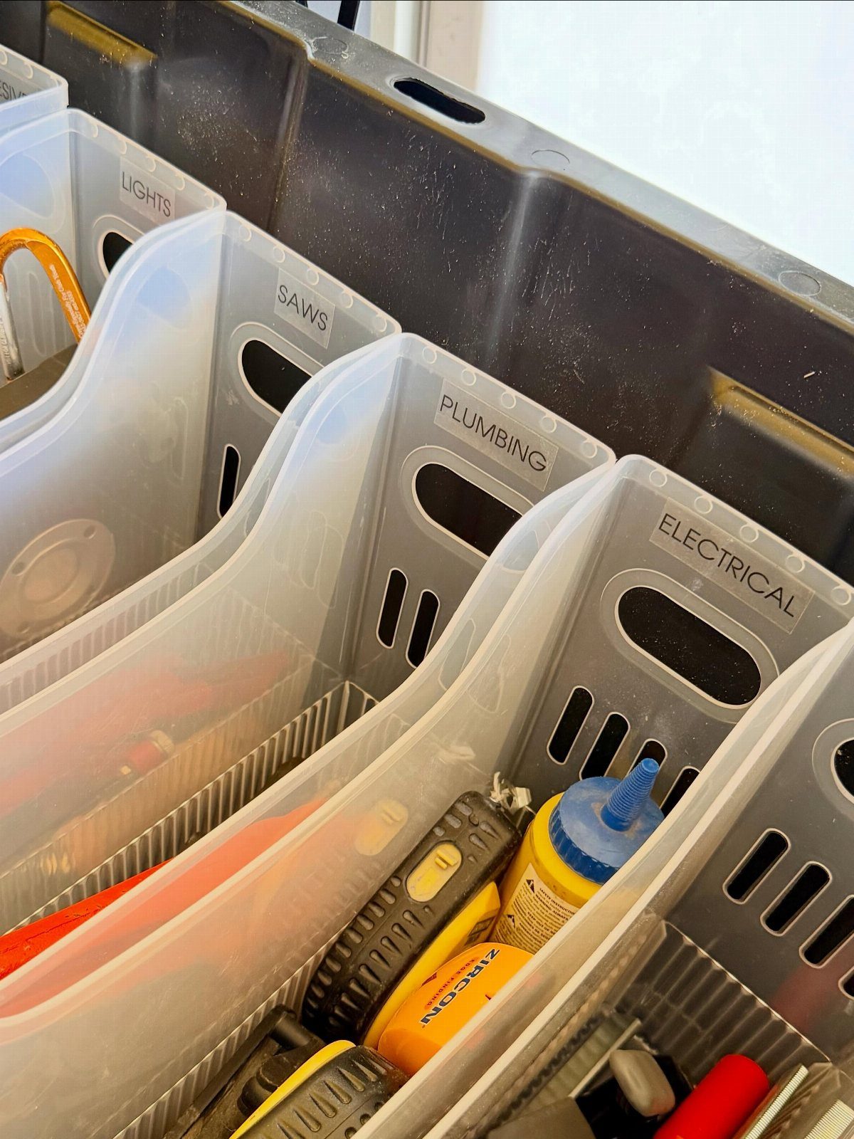 Translucent plastic storage bins labeled for saws, plumbing, and electrical tools inside a black container.