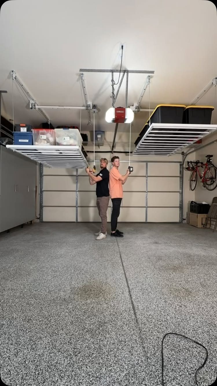 Two men operating ceiling-mounted overhead garage storage racks holding plastic bins with pulley ropes.