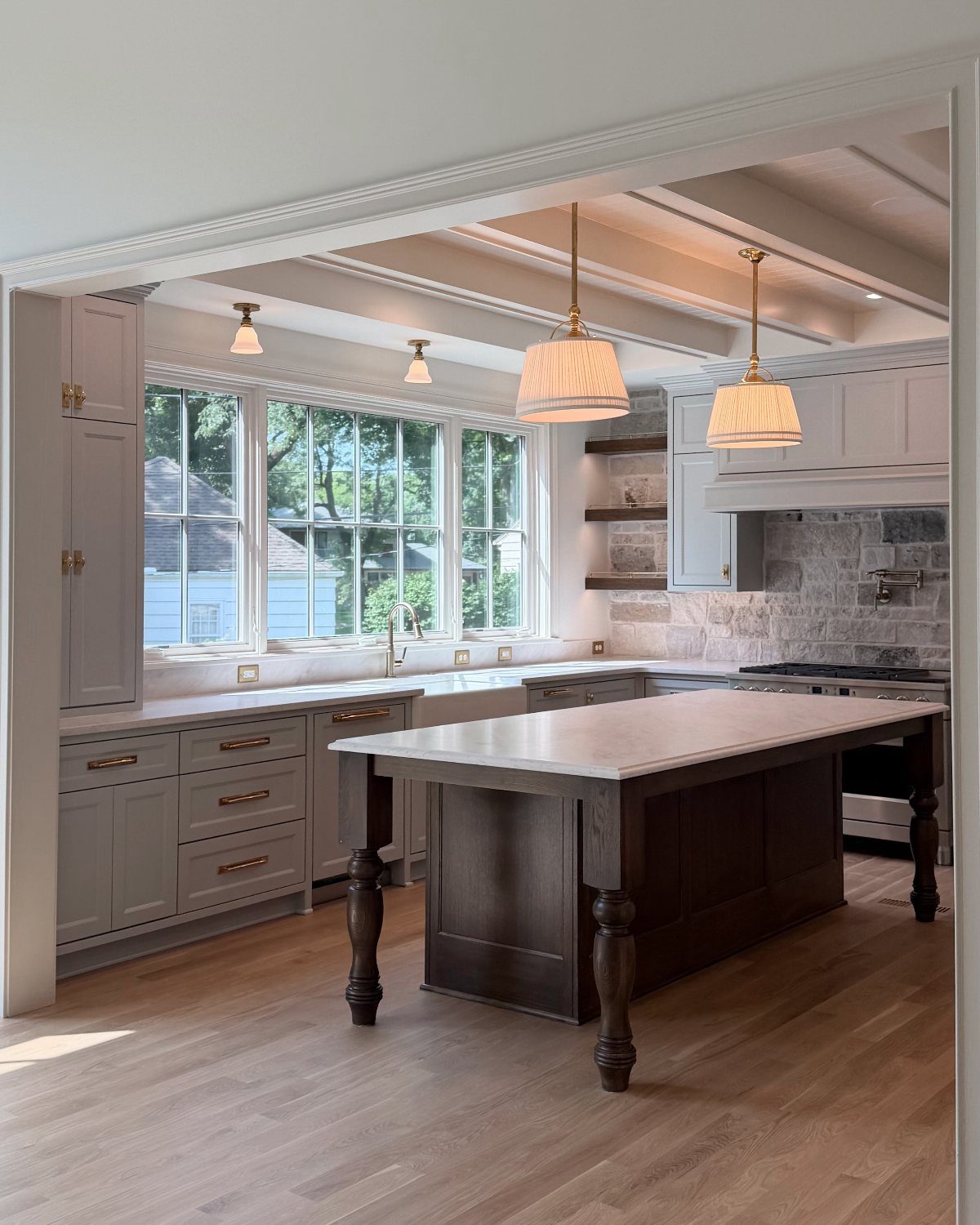 A bright farmhouse kitchen with grey-blue cabinets, a dark wood island, and large windows overlooking trees.