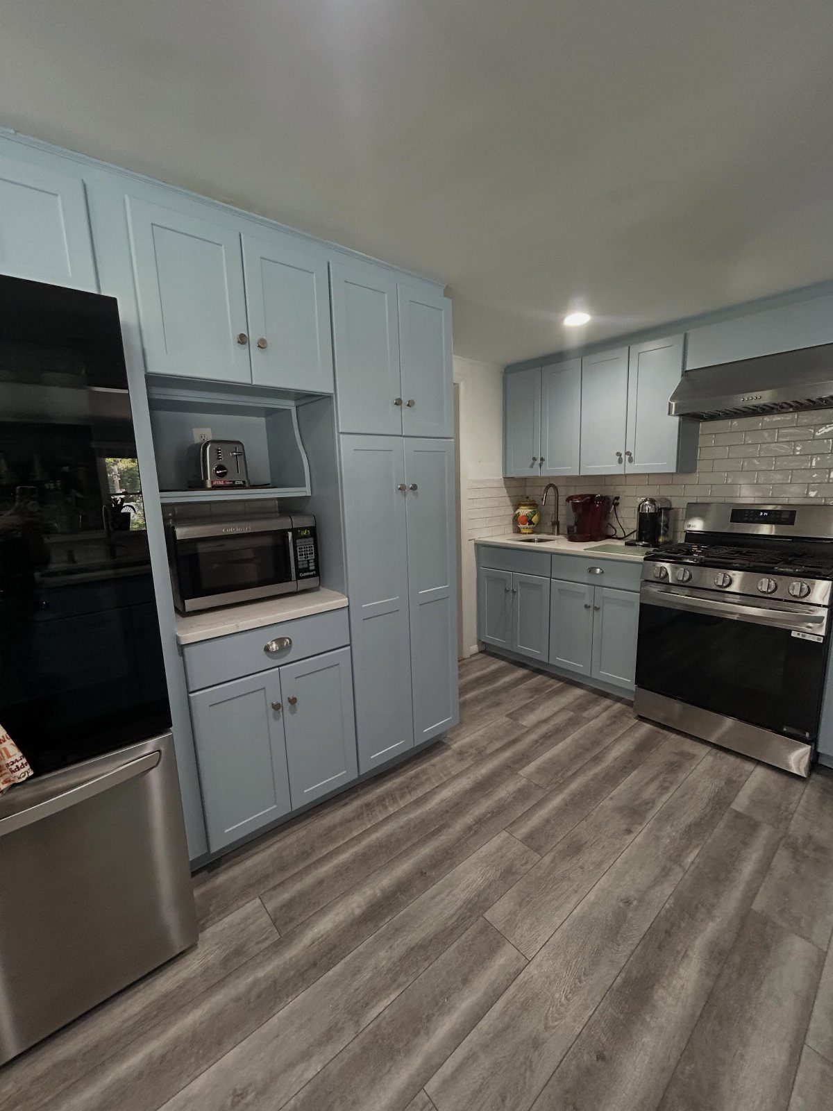 Light blue kitchen cabinets arranged with stainless steel appliances and grey wood-style flooring.