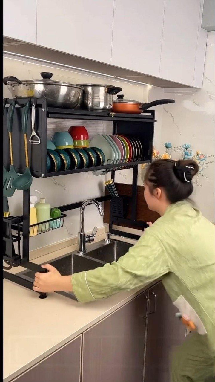 A person adjusting a black multi-tier metal organizer rack over a kitchen sink.