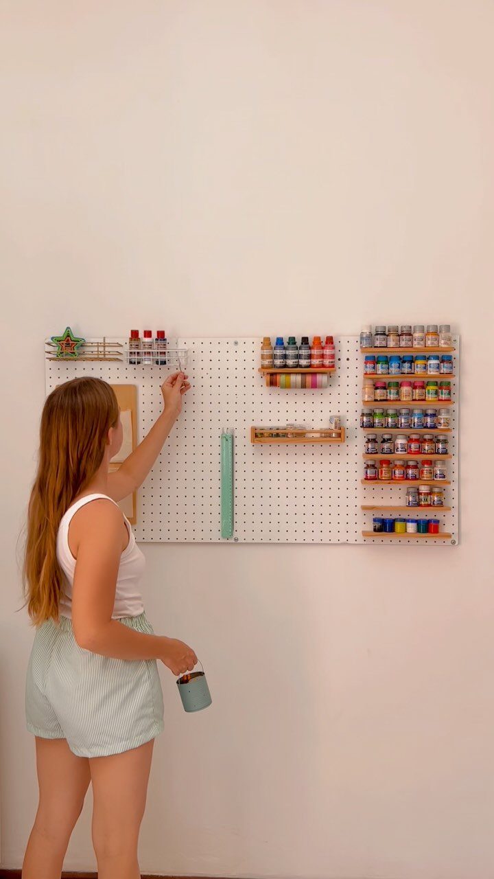 A woman organizing colorful paint bottles on a white wall-mounted pegboard system.