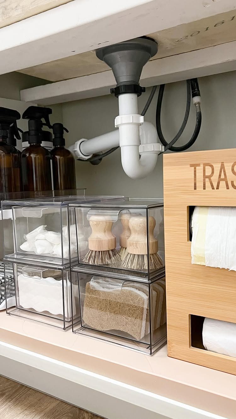 Clear plastic bins with sponges and wooden brushes organized under a white sink with plumbing pipes.