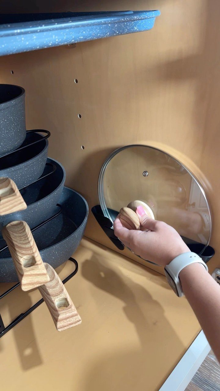 A child's hand reaching for a glass lid inside an organized kitchen cabinet with stacked pots.