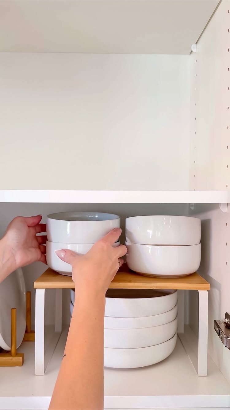 Hands placing white ceramic bowls onto a wooden shelf riser inside a white kitchen cabinet.