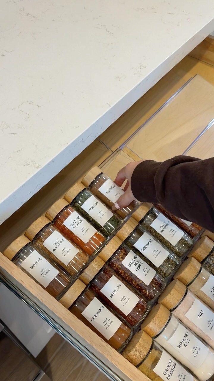 A hand reaching into a kitchen drawer filled with neatly organized glass spice jars.