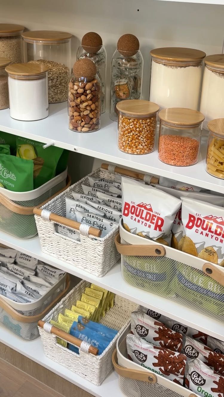 Glass jars with wooden lids and white baskets filled with snacks on white pantry shelves.