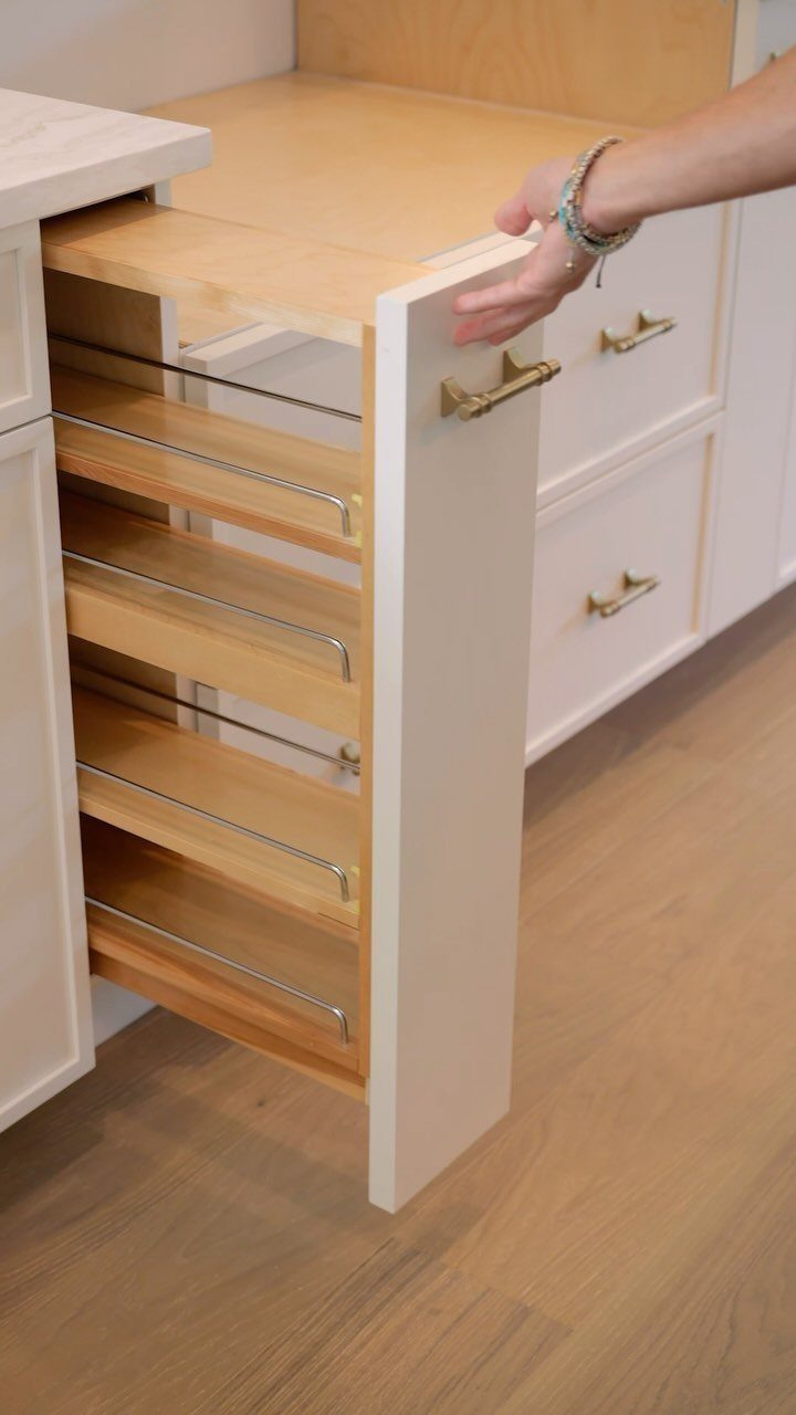 A hand reaches toward an open white kitchen cabinet containing several wooden pull-out shelves.