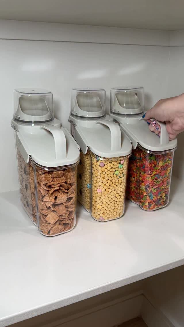 Three clear plastic cereal dispensers filled with colorful breakfast cereals sitting on a white kitchen shelf.