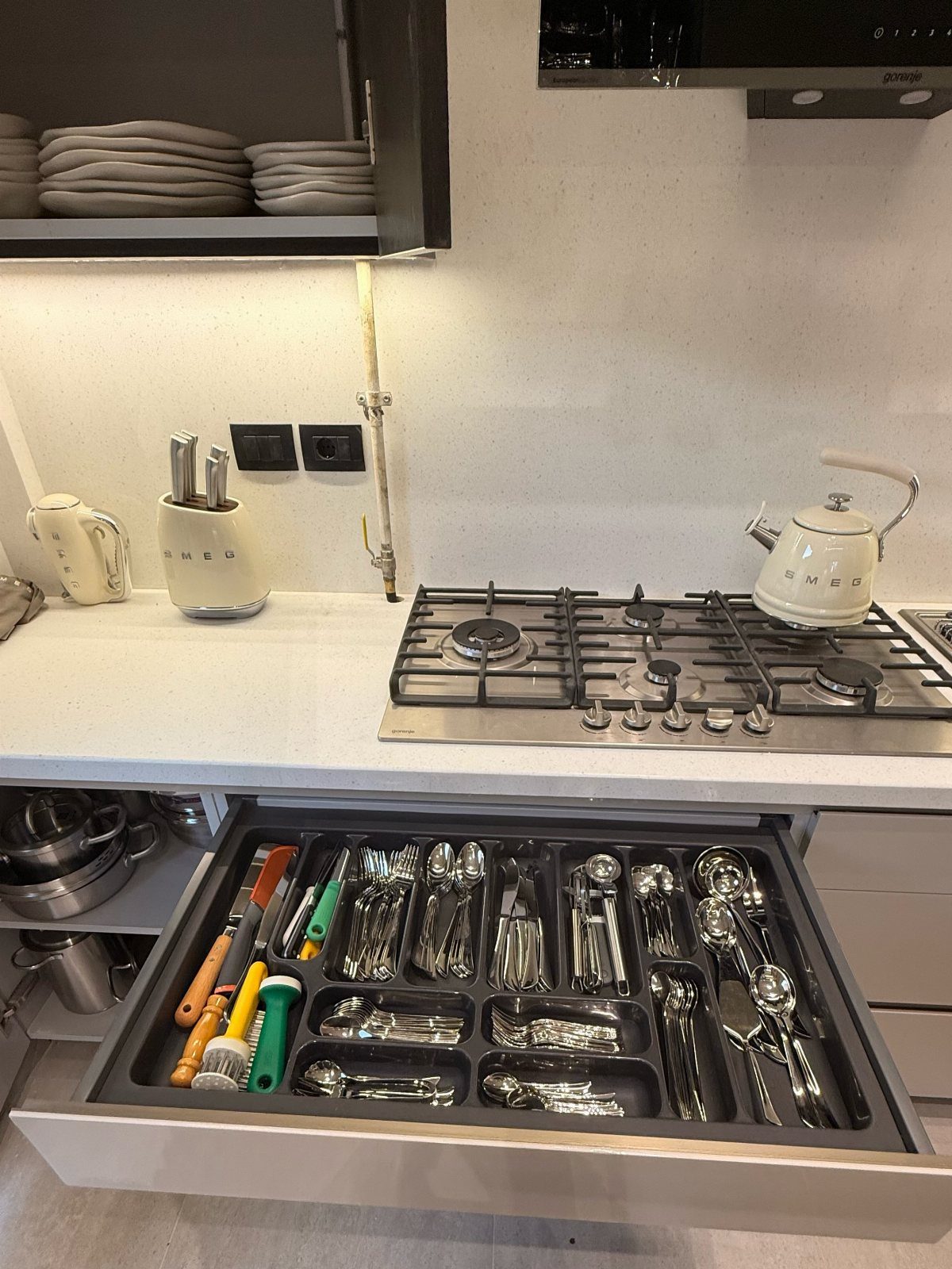 An open kitchen drawer filled with organized silver utensils and colorful kitchen tools in a grey tray.