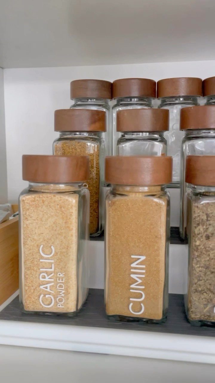 Glass spice jars with wooden lids and white labels arranged neatly on a white kitchen shelf.
