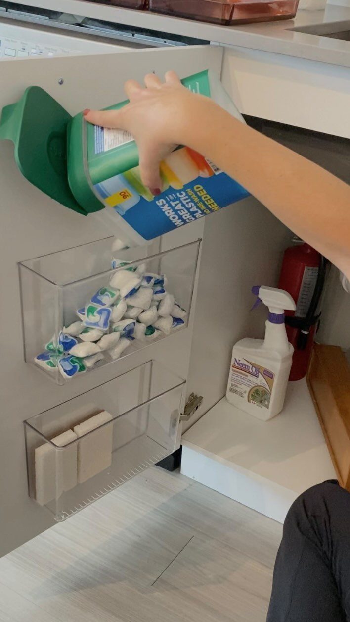A hand reaches for a plastic wrap box mounted on the inside of a white cupboard door.