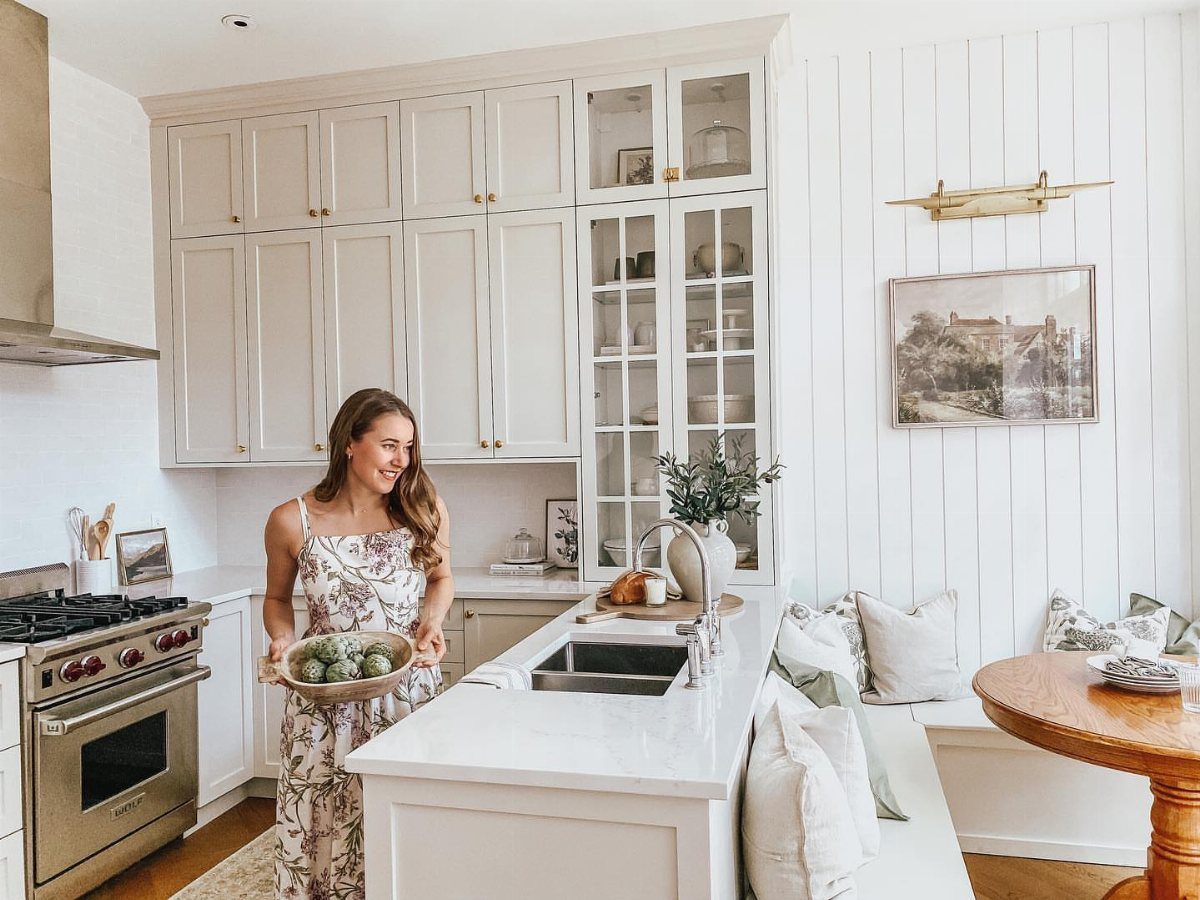 Woman in a floral dress standing in a bright kitchen with tall white cabinets.