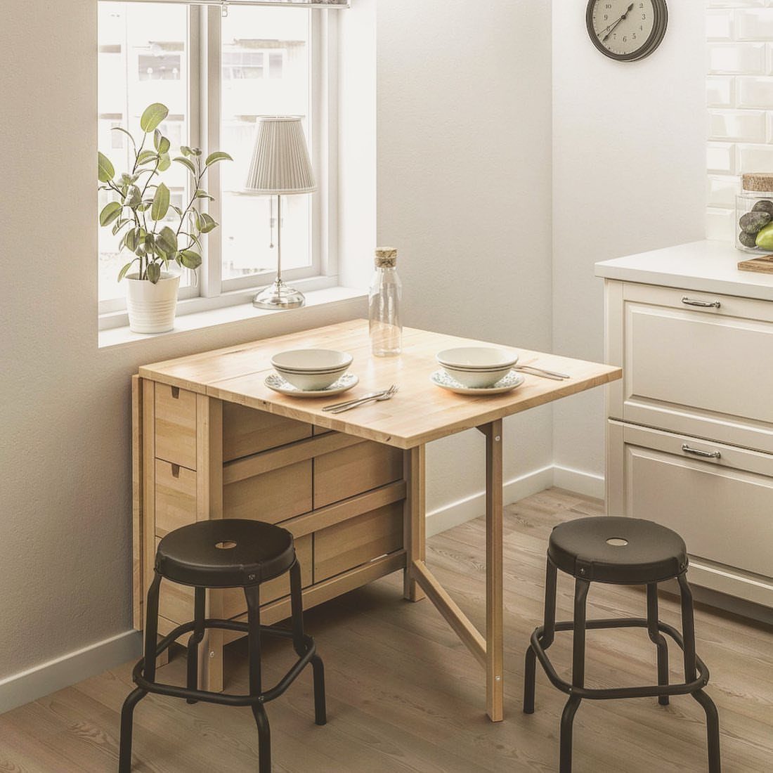 A light wood gateleg table set for two with black stools in a bright kitchen.