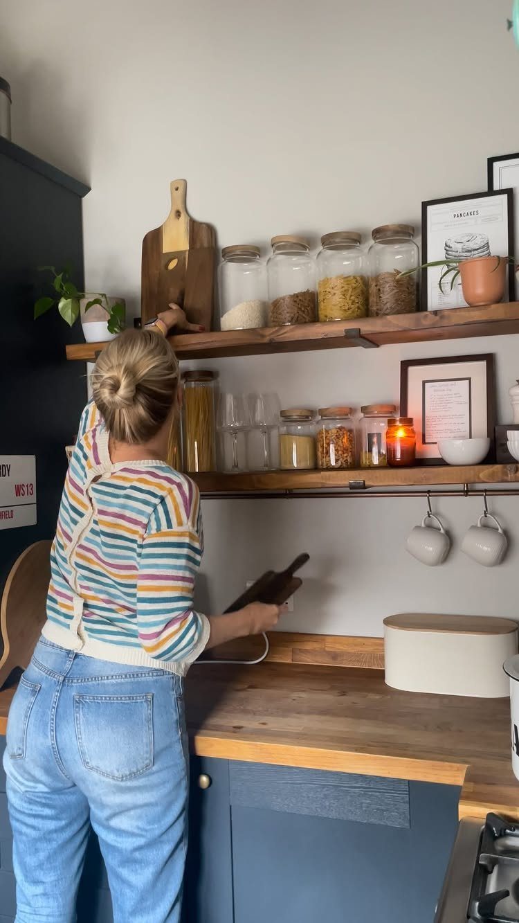 A person reaching for a wooden cutting board on dark wood open kitchen shelves.