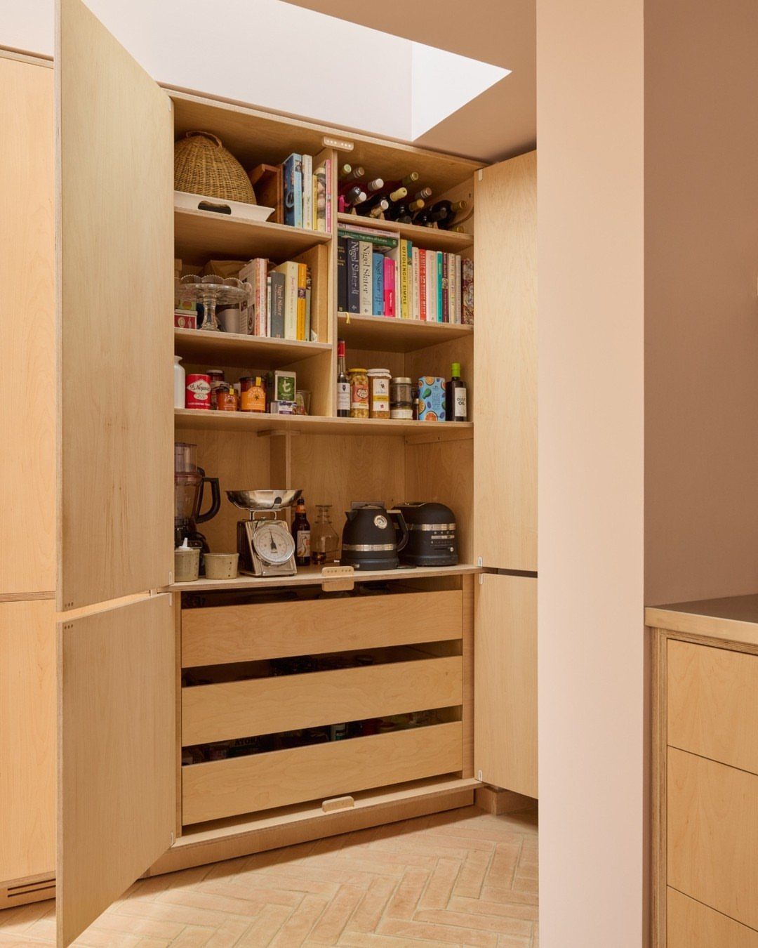 Light wood pantry cabinet with open shelves containing books, bottles, and various kitchen appliances.