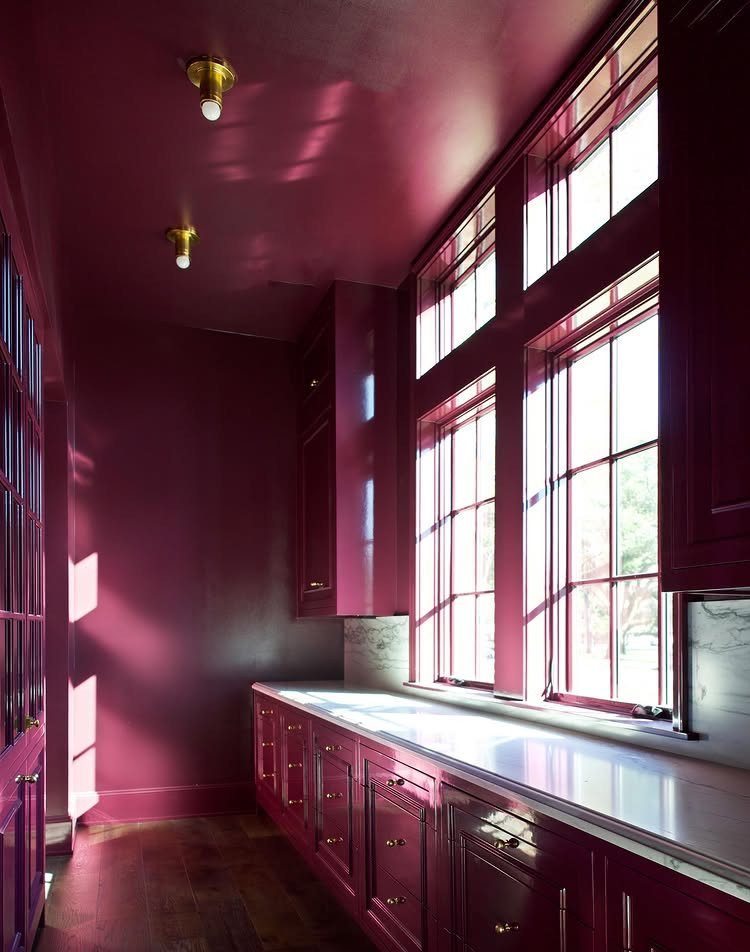 Deep berry colored kitchen cabinets and walls with white marble countertops and large windows.