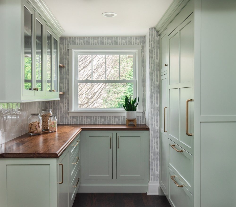 Mint green kitchen cabinets with wood countertops and brass handles near a window.