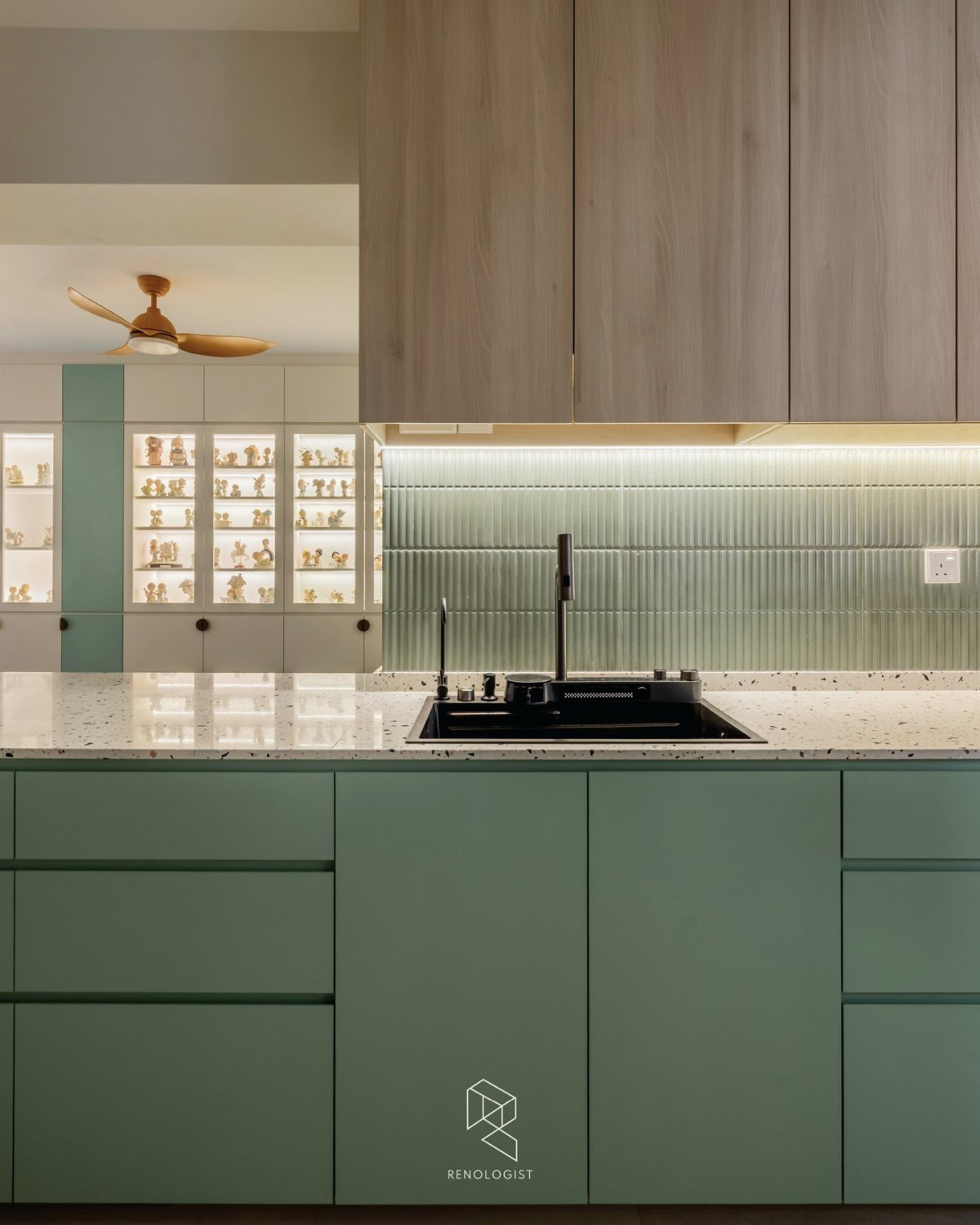 Sage green kitchen cabinets with a black sink and white speckled countertop under warm lighting.