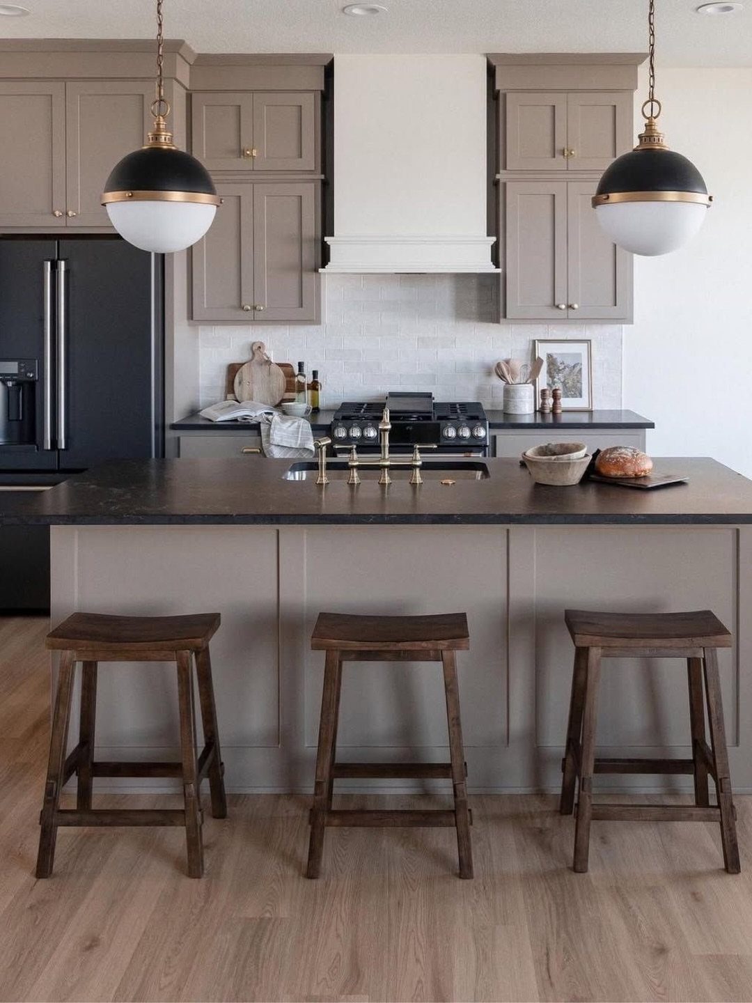 Taupe kitchen cabinets with a dark island, wooden stools, and black and gold pendant lights.