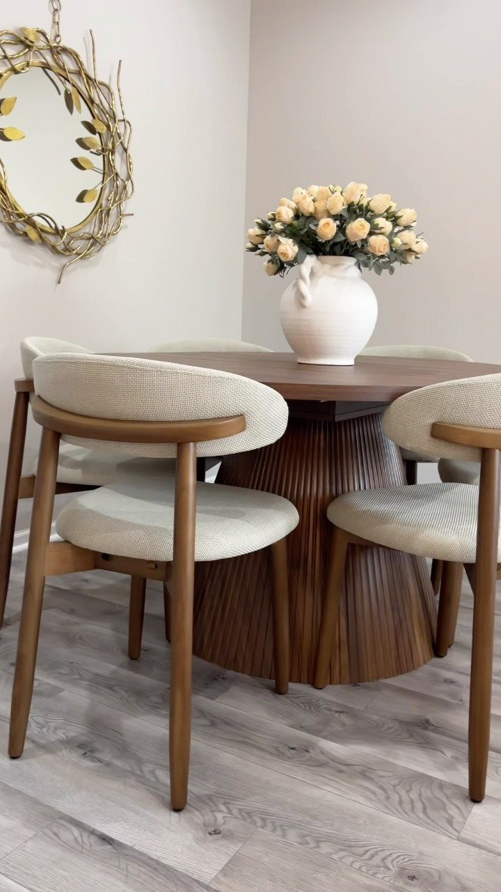 A round wooden dining table with cream roses and four upholstered chairs on light wood flooring.
